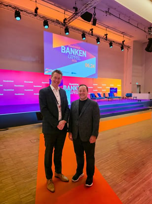 Zwei Personen im Sakko stehen vor der Bühne des Handelsblatt Banken Gipfels in Tönen von Orange, Blau, Lila und Rot.