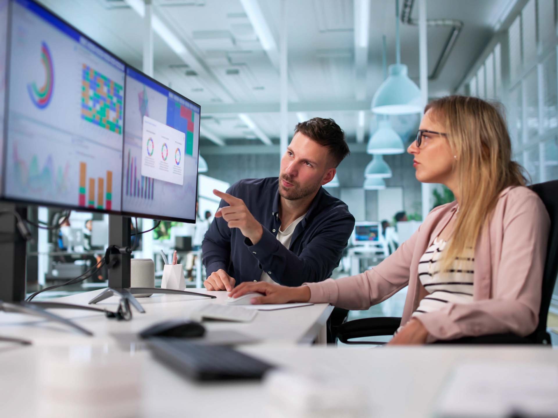 Eine Frau und ein Mann sitzen vor zwei Bildschirmen, die ein Dashboard zeigen, und besprechen Businessthemen