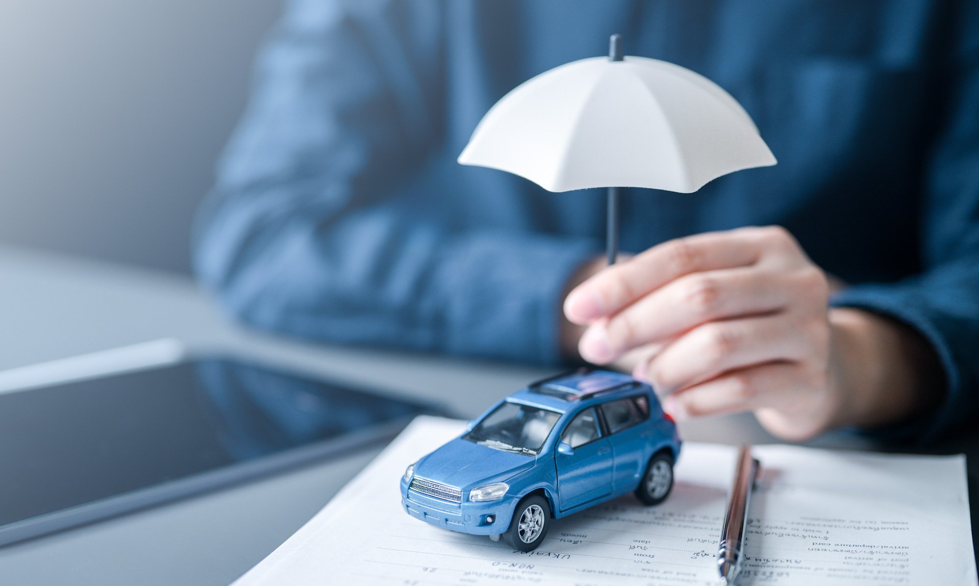 Weißer Schirm schützend in der Hand gehalten über einem blauen Spielzeugauto, dass auf Vertragsunterlagen steht